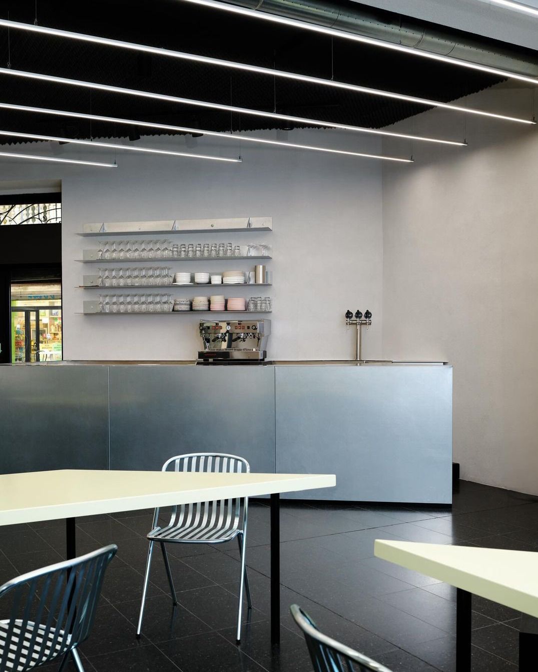 A futuristically conceived cocktail bar boasts sleek chrome furnishings, including steel chairs, bar counters, shelves, and fittings, alongside black tiling, lime green tables, and an edgy atmosphere.