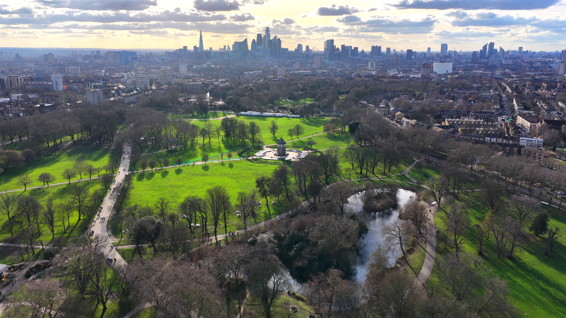 Victoria Park in East London with the central London skyline visible in the background. The image shows green parkland, tree-lined paths, and open spaces contrasting with modern city buildings