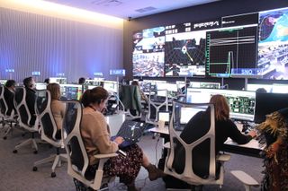 people sit at computers in front of a large wall of screens showing earth and various graphs