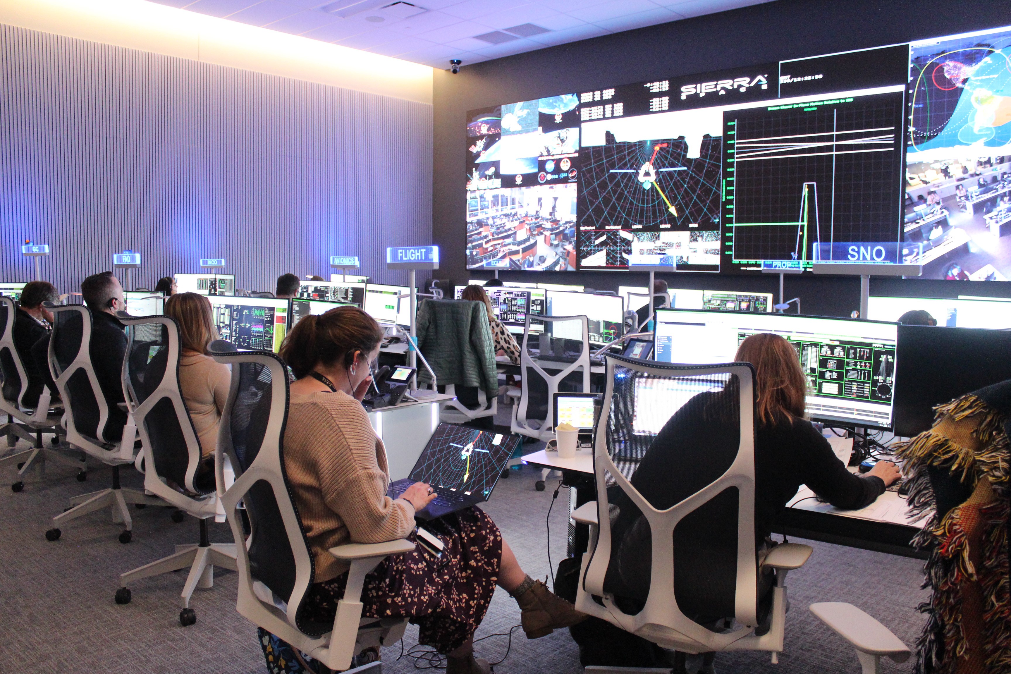 people sit at computers in front of a large wall of screens showing earth and various graphs