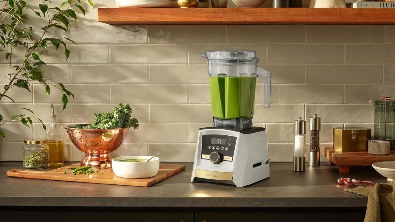 A Vitamix A3500 Ascent Series Smart Blende in nWhite with Gold Accents on a dark kitchen counte, next to a wooden cutting board and copper colander and green house plants