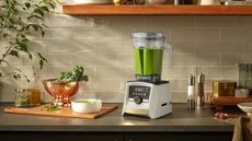 A Vitamix A3500 Ascent Series Smart Blende in nWhite with Gold Accents on a dark kitchen counte, next to a wooden cutting board and copper colander and green house plants