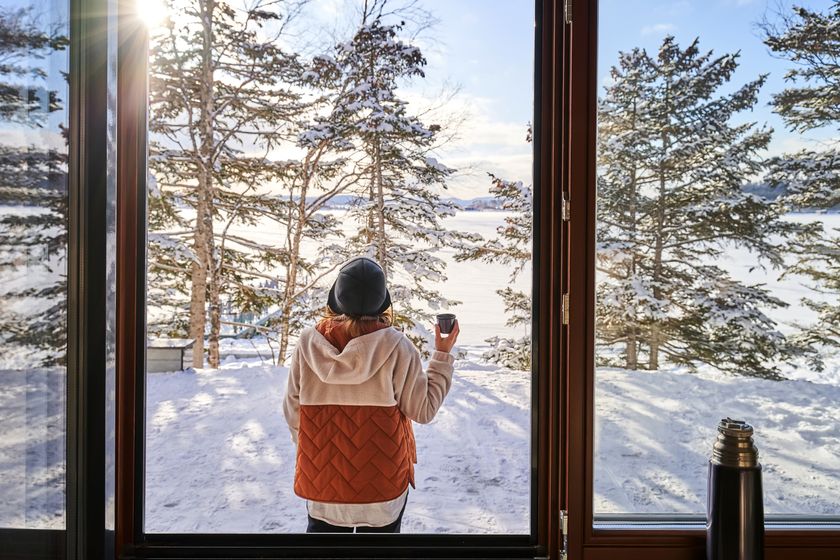 woman standing outside a window looking at a snowy scene holding a tea