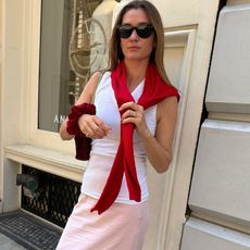 French style influencer Anne-Laure Mais poses on a NYC sidewalk wearing Ray-Ban sunglasses, a red sweater on her shoulders, white tank top, red scrunchie, and a light-pink skirt