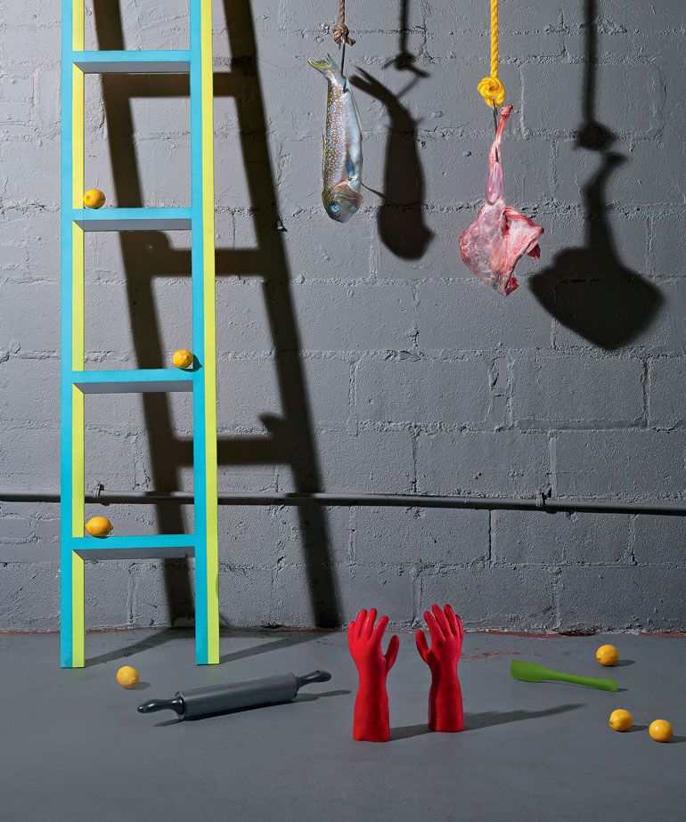 meat hanging against a grey background and floor with coloured ladder to the left, red gloves, fruit and kitchen utensils on the floor