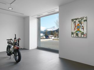 Installation view featuring one electric motorcycle by a window in a room with grey flooring and white walls. There is a colourful piece of art on the wall