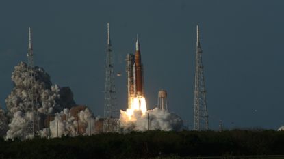 An orange and white rocket blasts off a launchpad, billowing gray smoke behind it