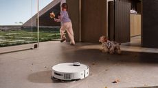 A white circular Dreame X50 Ultra vacuuming a floor with brown leaves, with a child and a dog running into the garden