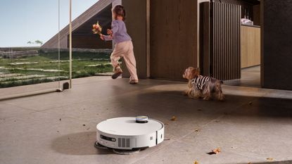 A white circular Dreame X50 Ultra vacuuming a floor with brown leaves, with a child and a dog running into the garden