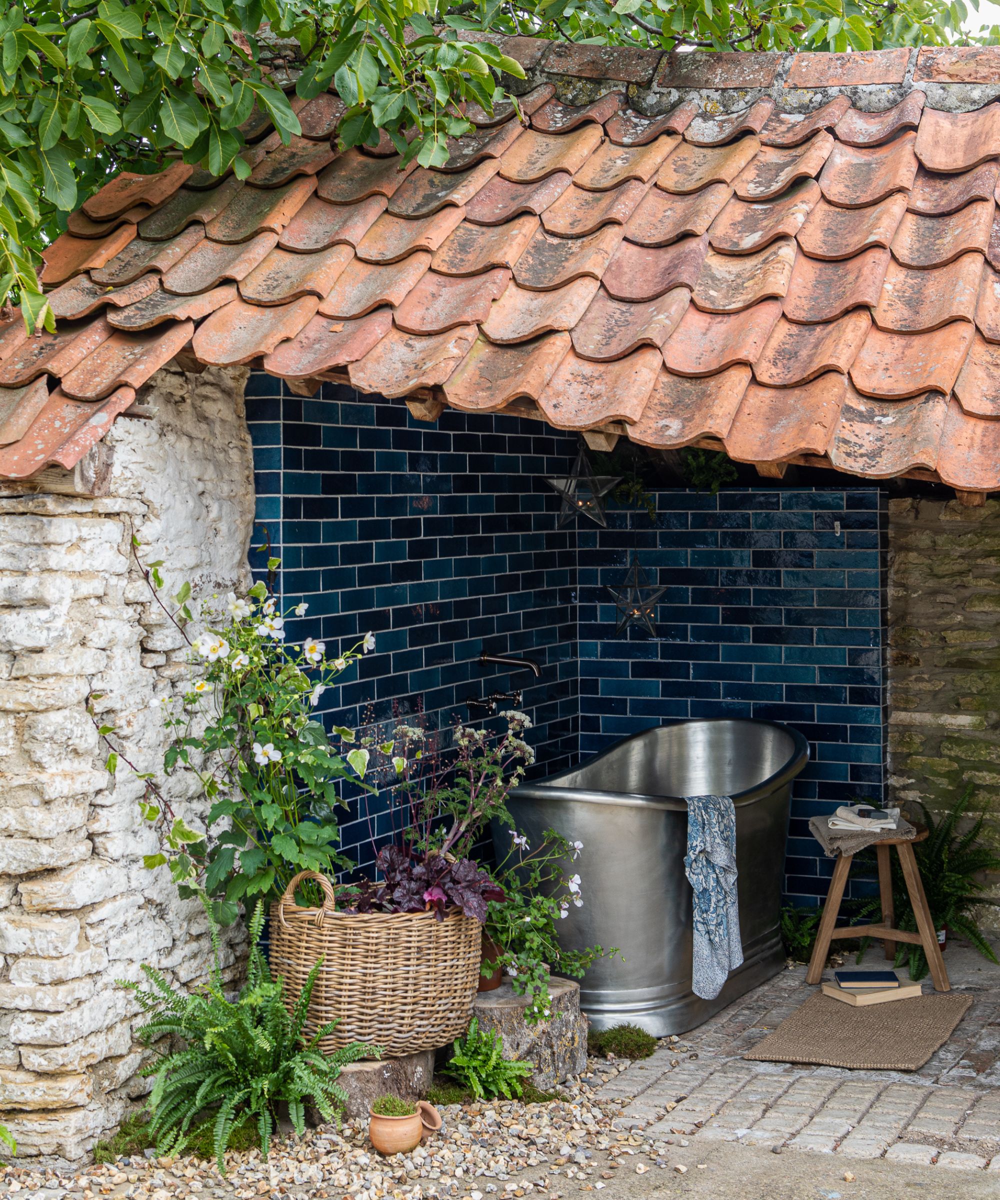 A rustic outdoor bathing nook tucked under a terracotta roof overhang, with deep navy zellige brick tiles, a freestanding pewter metal tub, wicker basket of plants and gravel floor.