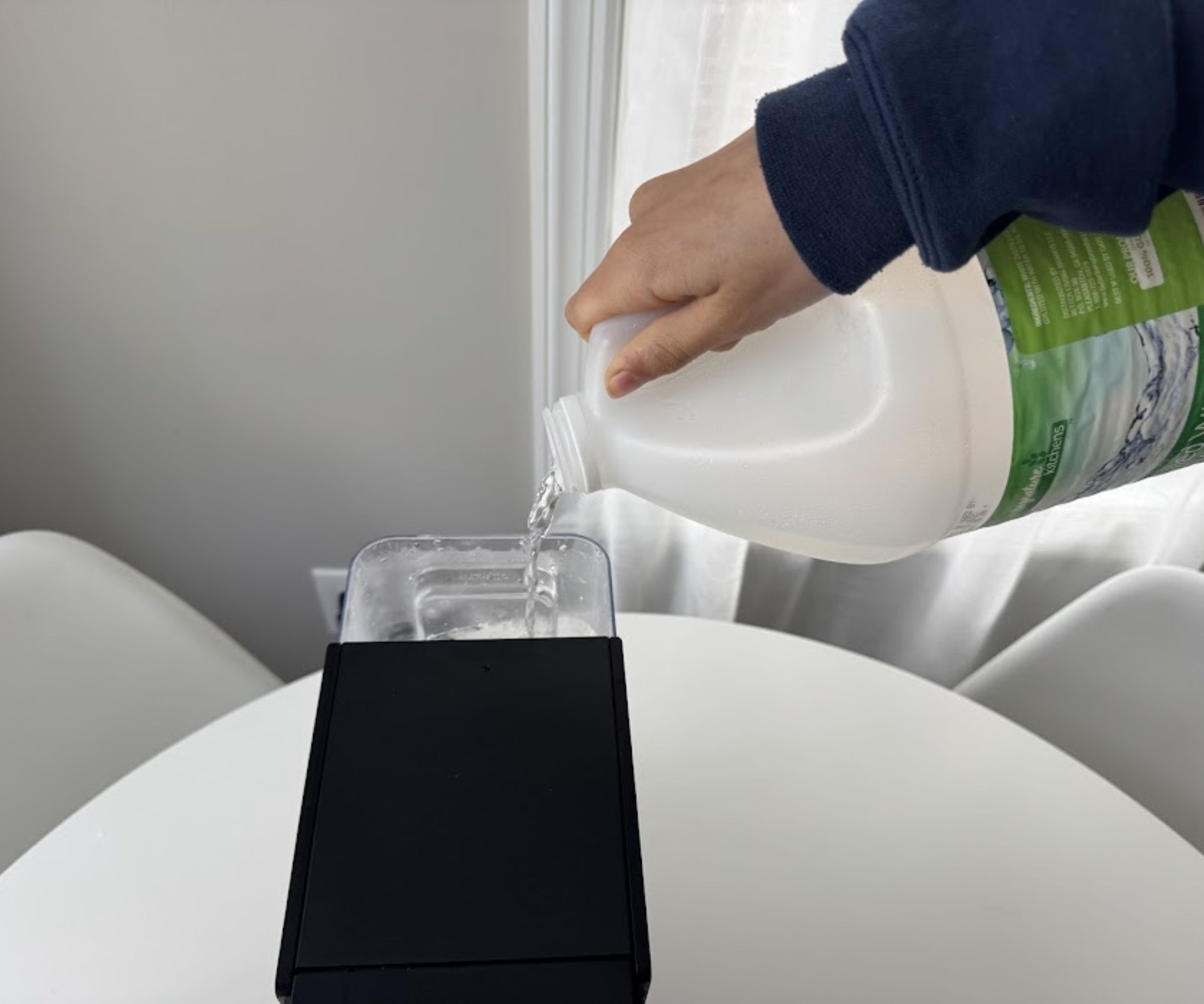 A hand pouring white vinegar from a large plastic bottle into the back reservoir of a black Keurig coffee maker.