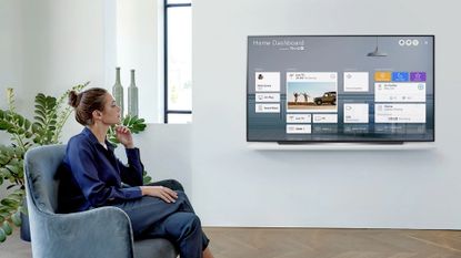 woman watching the LG 77 Class CX Series OLED 4K UHD Smart webOS TV, one of the best 75 inch TV options, in a white living room with a window, grey vases and a plant behind her