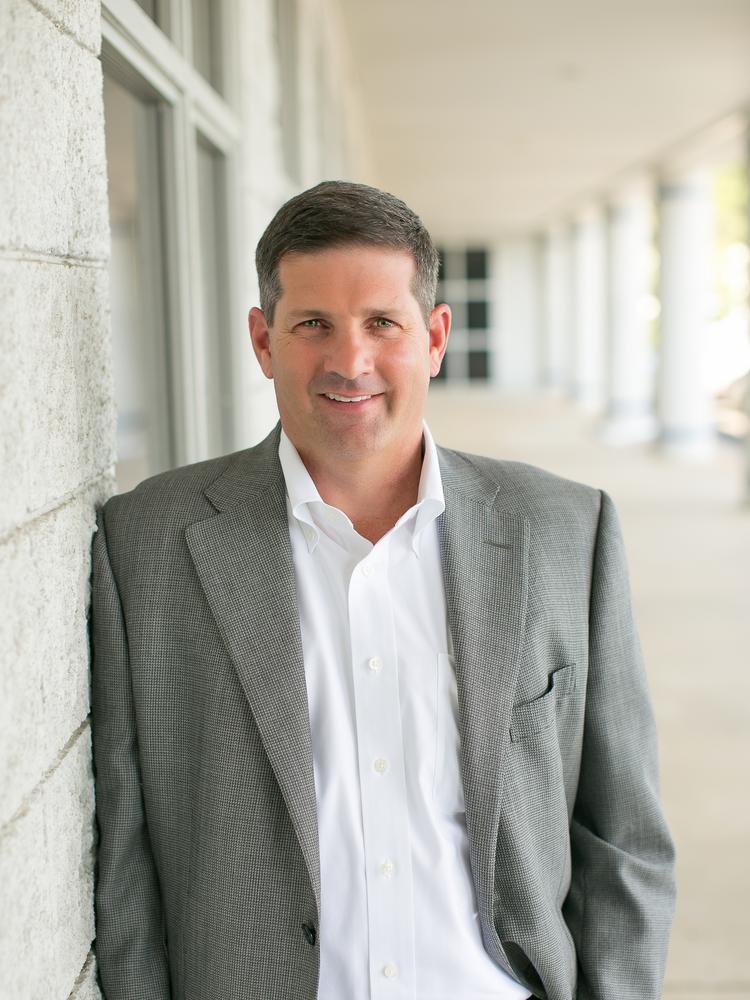 Photograph of Jimmy Hiller wearing white shirt and gray suit jacket, leaning on white bricked wall with blurred outdoor background.