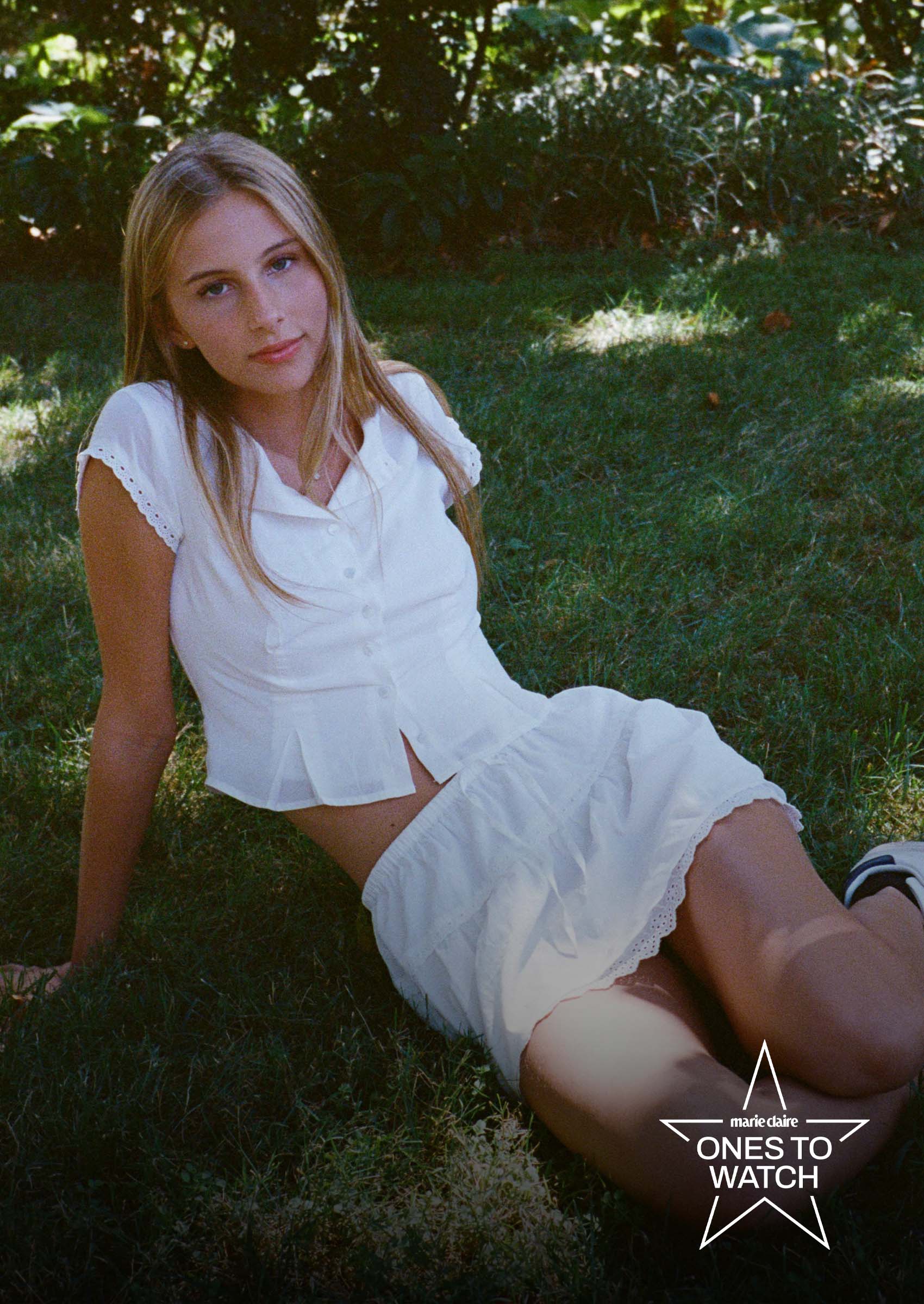 singer romy mars poses lying down in the grass wearing a white blouse and white skirt