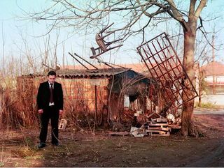 A dark haired man in a black suit stands with head down in front of a ramshackle garage with an old dining chair strung up above the roof and an old iron bedstead hanging vertically to one side.