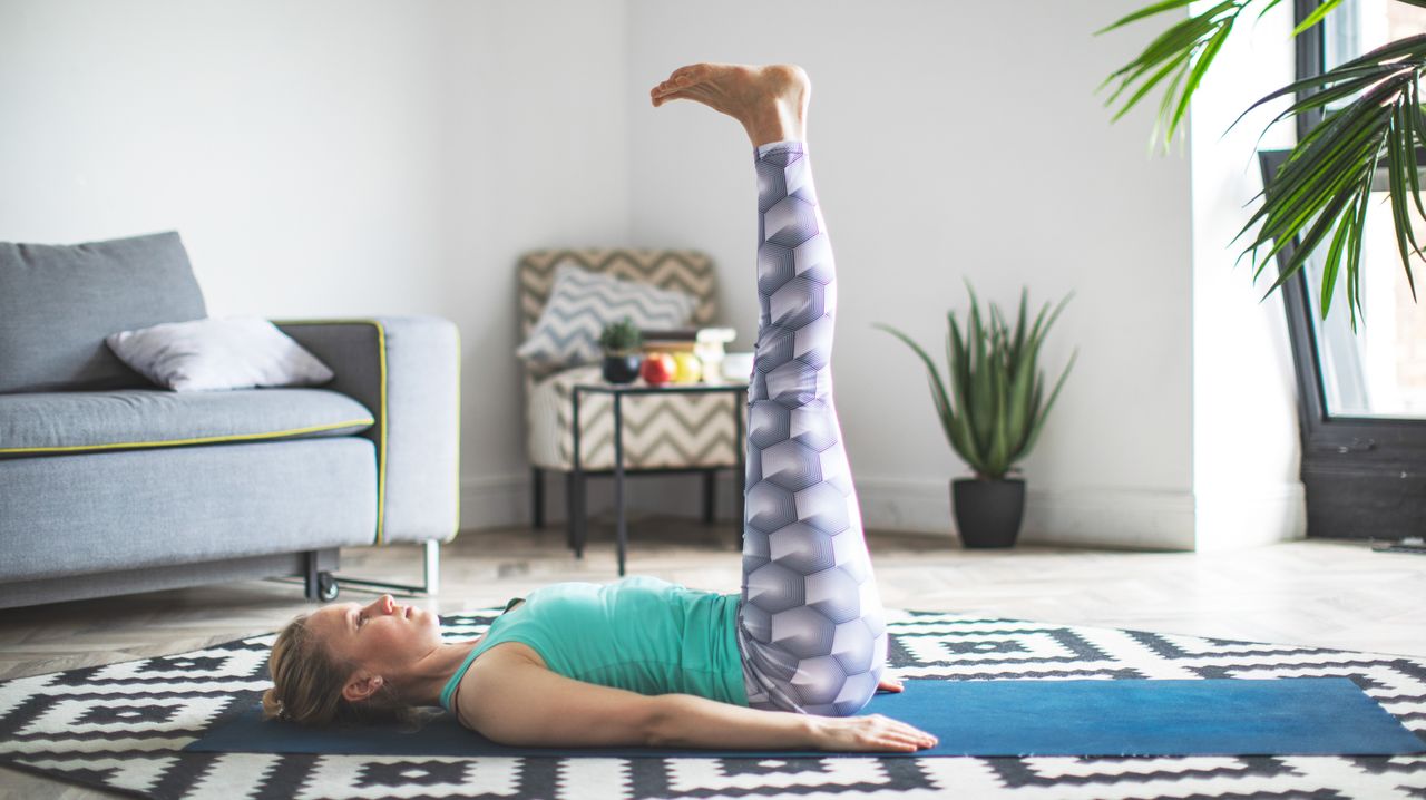 A woman practices Pilates at home in a living room on her mat. She is on her back, arms by her sides, with her legs straight and feet in the air. Behind her is a couch, plants and glass table.