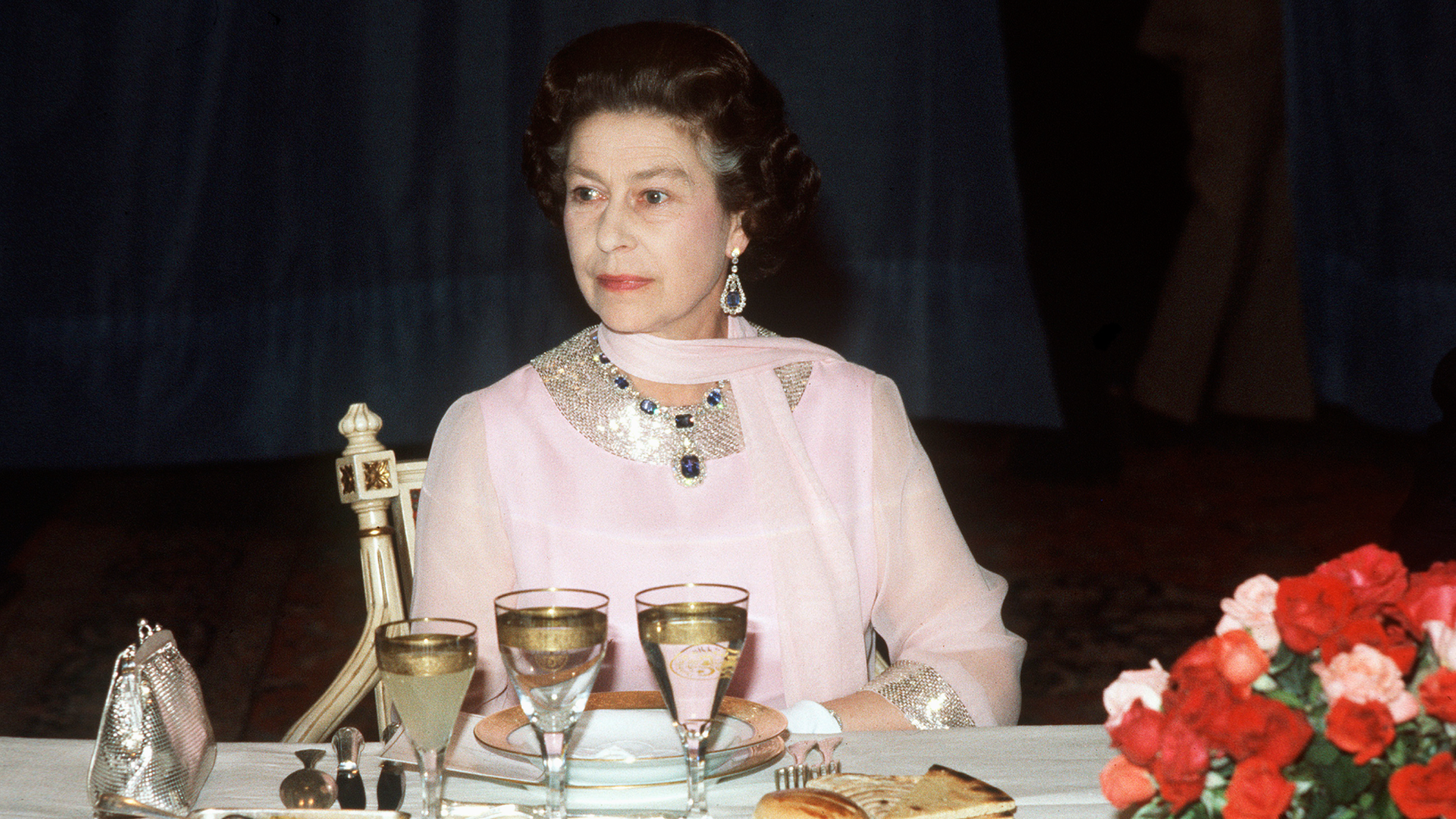 Queen Elizabeth wearing a pink dress sitting at a dining table
