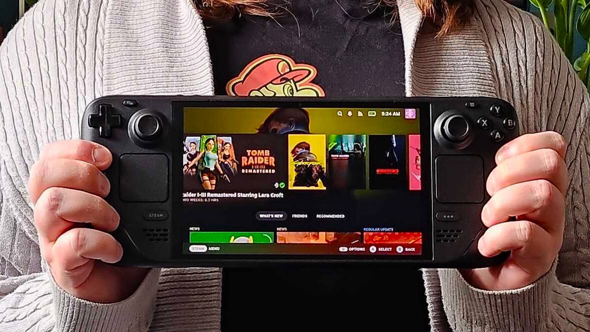 Hands holding Steam Deck OLED with SteamOS main screen displayed.
