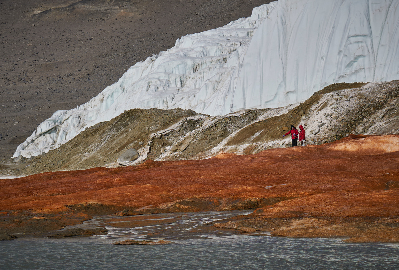 Two researchers stand on red-stained sediments near Blood Falls in Antarctica.