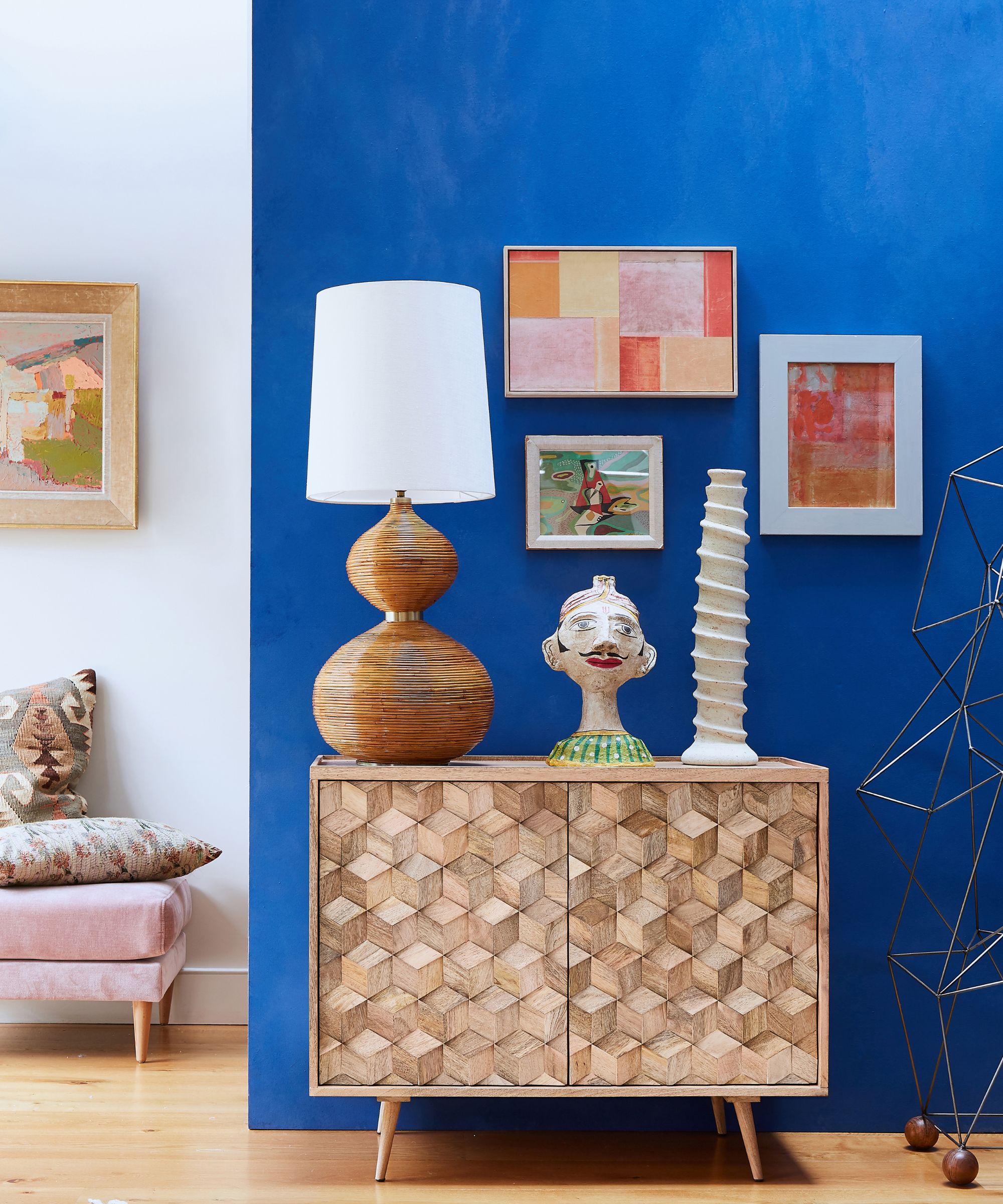 A bold blue feature wall behind a wooden sideboard. Sculptural objects, a table lamp and framed abstract art sit against the colour. Pale flooring and lighter furniture nearby balance the strong wall, keeping the space bright and visually striking overall.