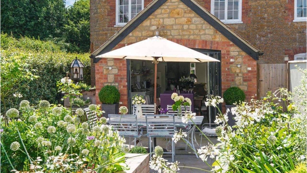 Cottage exterior wall with a large open door into the garden, patio seating area with table and chairs and plants in large wooden raised beds.