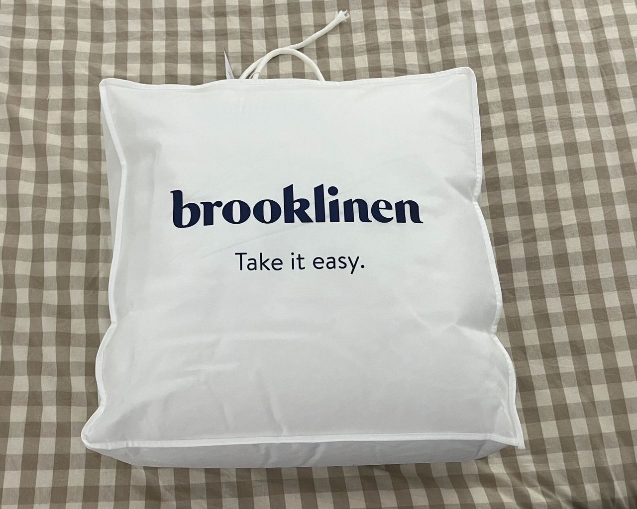 A Brooklinen down alternative duvet in a white bag with dark navy lettering, pictured on pale brown and white gingham bedding