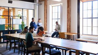 A diverse mix of workers in a co-working space, which features a large meeting table and large bright windows. The space is largely empty.