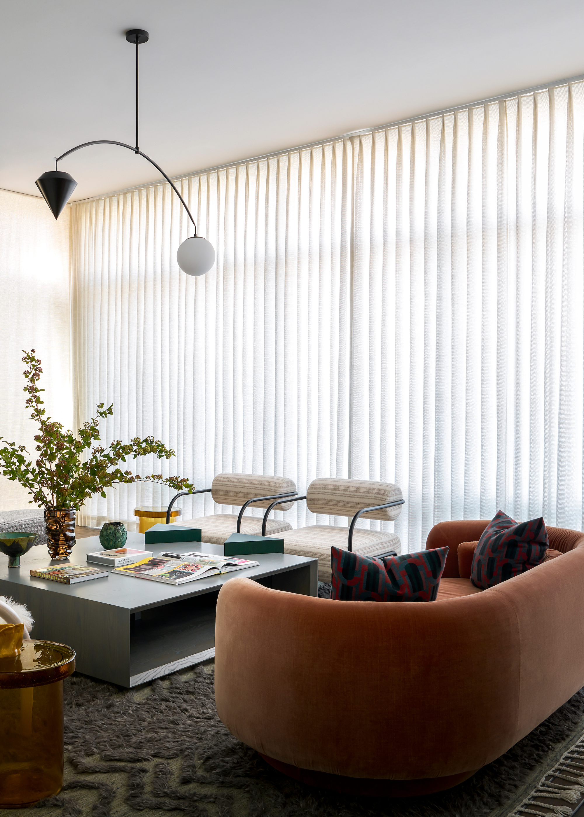 A bright living room with a rounded soft orange sofa, a pair of curved armchairs with cylindrical white cushions, an amber glass side table, a rectangular storage coffee table with an open book