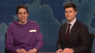 Pete Davidson in a purple sweatshirt on the set of Weekend Update alongside Colin Jost in a gray suit on Saturday Night Live. 