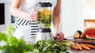 Blender full of fruit and vegetables on a kitchen countertop surrounded by more fruit and veg