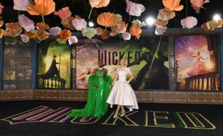 Cynthia Erivo in a bright green gown and Ariana Grande in a pale pink dress standing on a black red carpet reading WICKED with a movie poster behind them