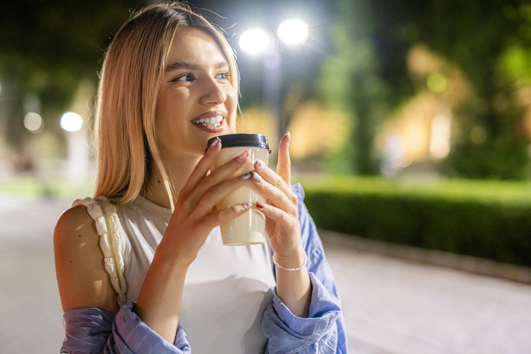 A woman talks along a street in the evening holding a disposable coffee cup with both hands.
