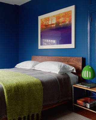 A bedroom painted in a blue Benjamin Moore paint with a purple framed poster on the wall, a green throw on the bed and a neon green table lamp on the bedside table