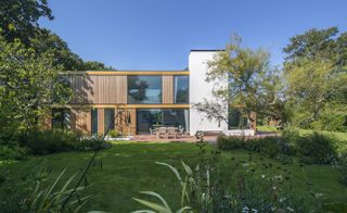 Woodpeckers by Ström Architects, grass lawn, plants and shrubs, wooden two story building, with windows and white tower section at the right handside, trees to the left and right, clear blue sky