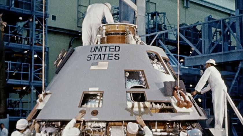 technicans in white coveralls work above, beside and below a space capsule