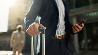 A businessman walking down a street with a suitcase and mobile phone in hand.