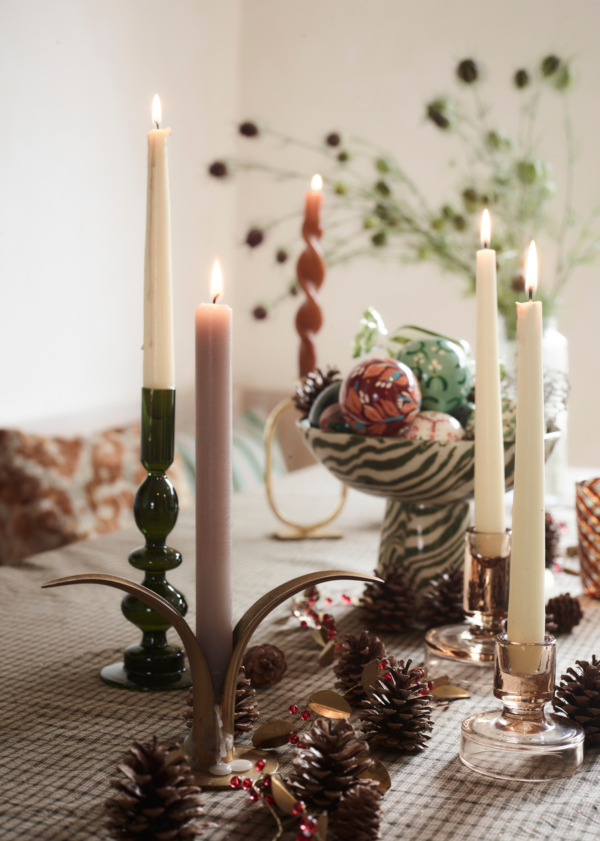 A festive table display with lit taper candles, pinecones, berries and a bowl of baubles with thistles in a vase in the background