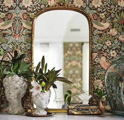 gilded mirror and grecian statue-style vases on a shelf with floral wallpaper behind