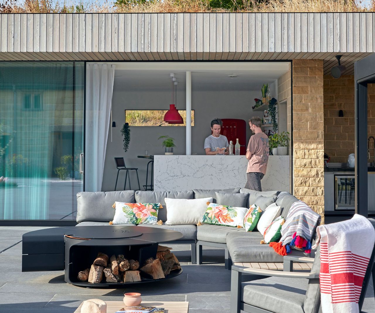 Two men at a marble-topped bar inside a timber-clad pool house with red pendant lights and a red Smeg fridge, with a large grey corner sofa and fire pit on the terrace outside.