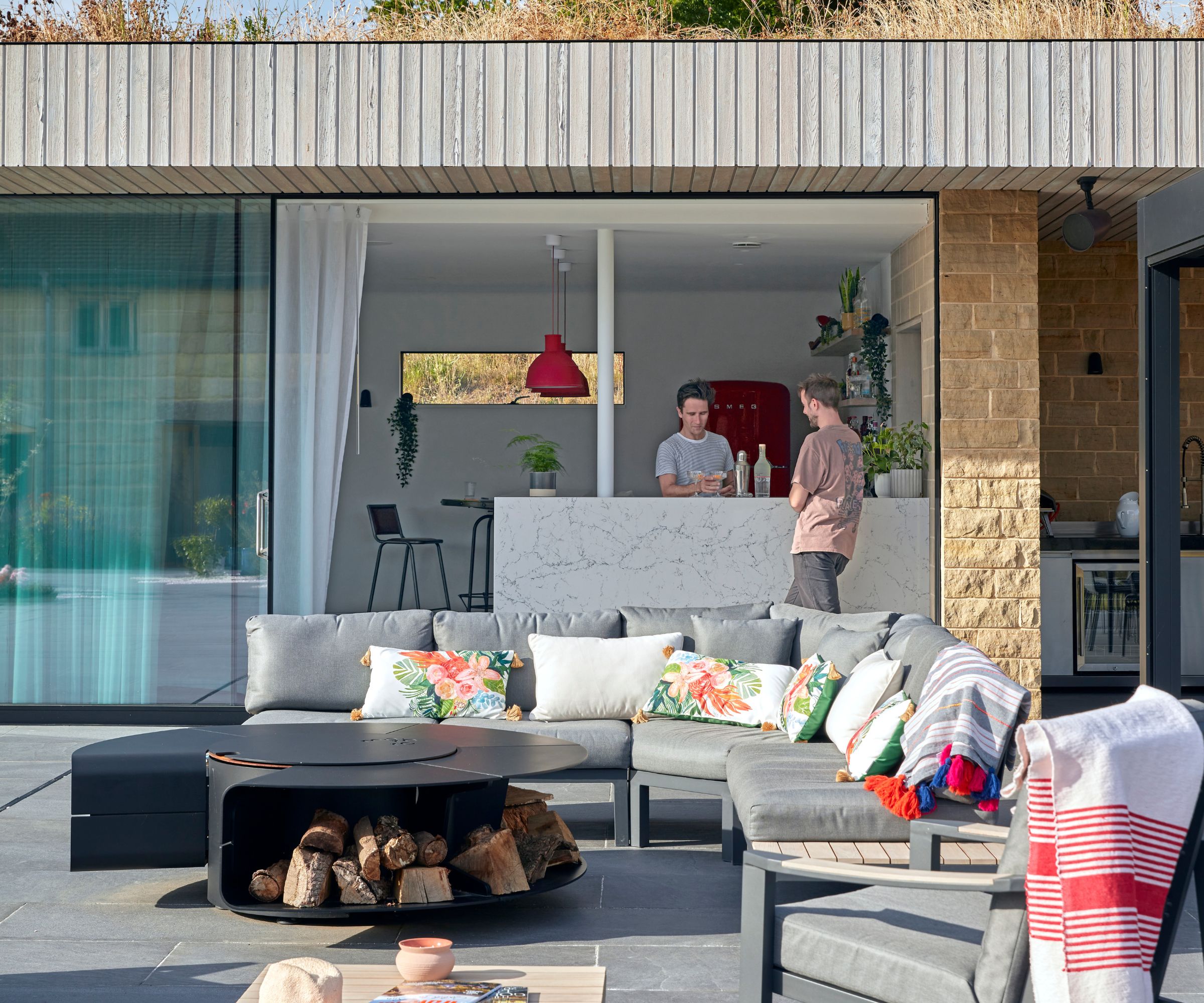 Two men at a marble-topped bar inside a timber-clad pool house with red pendant lights and a red Smeg fridge, with a large grey corner sofa and fire pit on the terrace outside.