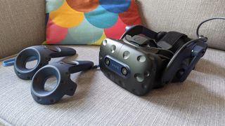 Ett HTC Vive Pro 2-headset ligger bredvid sina tillhörande kontroller i en grå soffa framför en färgglad kudde.