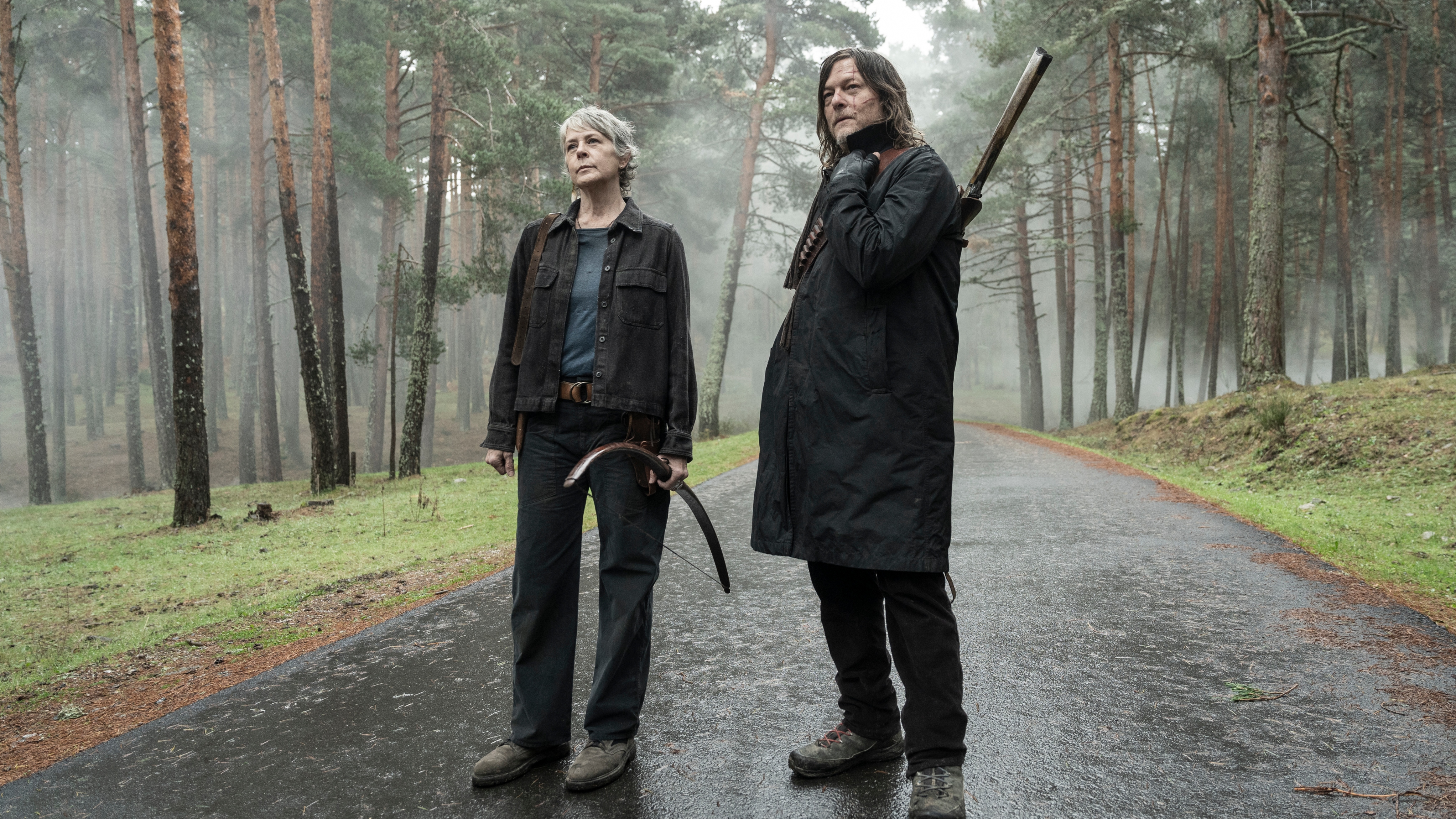 Melissa McBride as Carol Peletier and Norman Reedus as Daryl Dixon standing on a path in the woods. Carol is holding a bow in her hand and Daryl has a gun on his back.