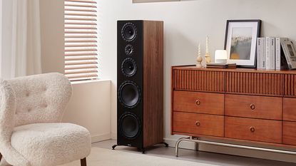 Elipson Prestige Facet II floorstanding speaker in the corner of a beige room. It's a walnut speaker with four drivers and a tweeter, with a black front panel. It's sitting next to a walnut sideboard with books, candles and a painting on top of the furniture.