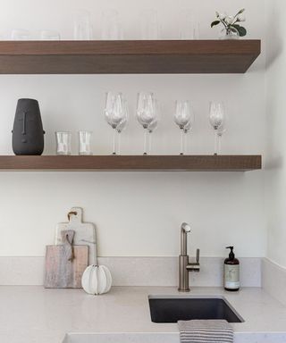 Neutral kitchen space focusing on two wooden shelves positioned above the kitchen sink. Styled in a minimalist way with glassware, decorative vases and plant. Natural, cream stone countertop with marble and wooden chopping boards, hand soap and silver tap