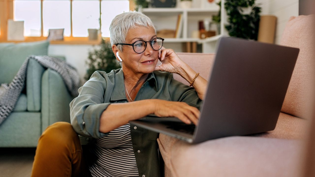 An older woman sits on the floor in front of her sofa and shops online on a laptop.