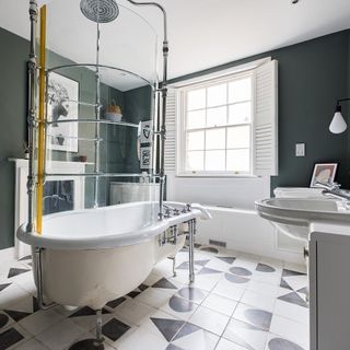 Roll-top bath with a curved screen on white tiles with black patterns next to fireplace and double vanity with bench seat under the shuttered windows