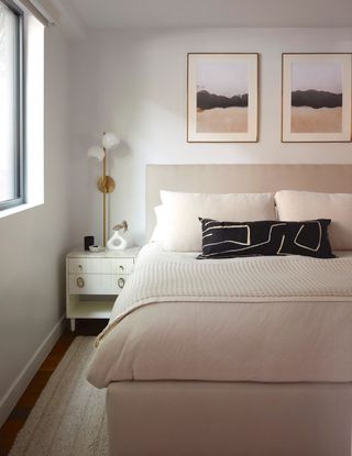 White bedroom space with bed dressed in neutral bedding and dark cushions, alongside minimally decorated nightstand with soft-edged trinketstand