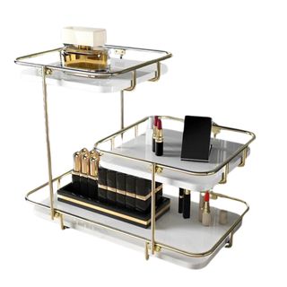 Three tiered gold metal and white shelved organizer with 3 staggered shelves. Perfumes and makeup items such as lipsticks are pictured