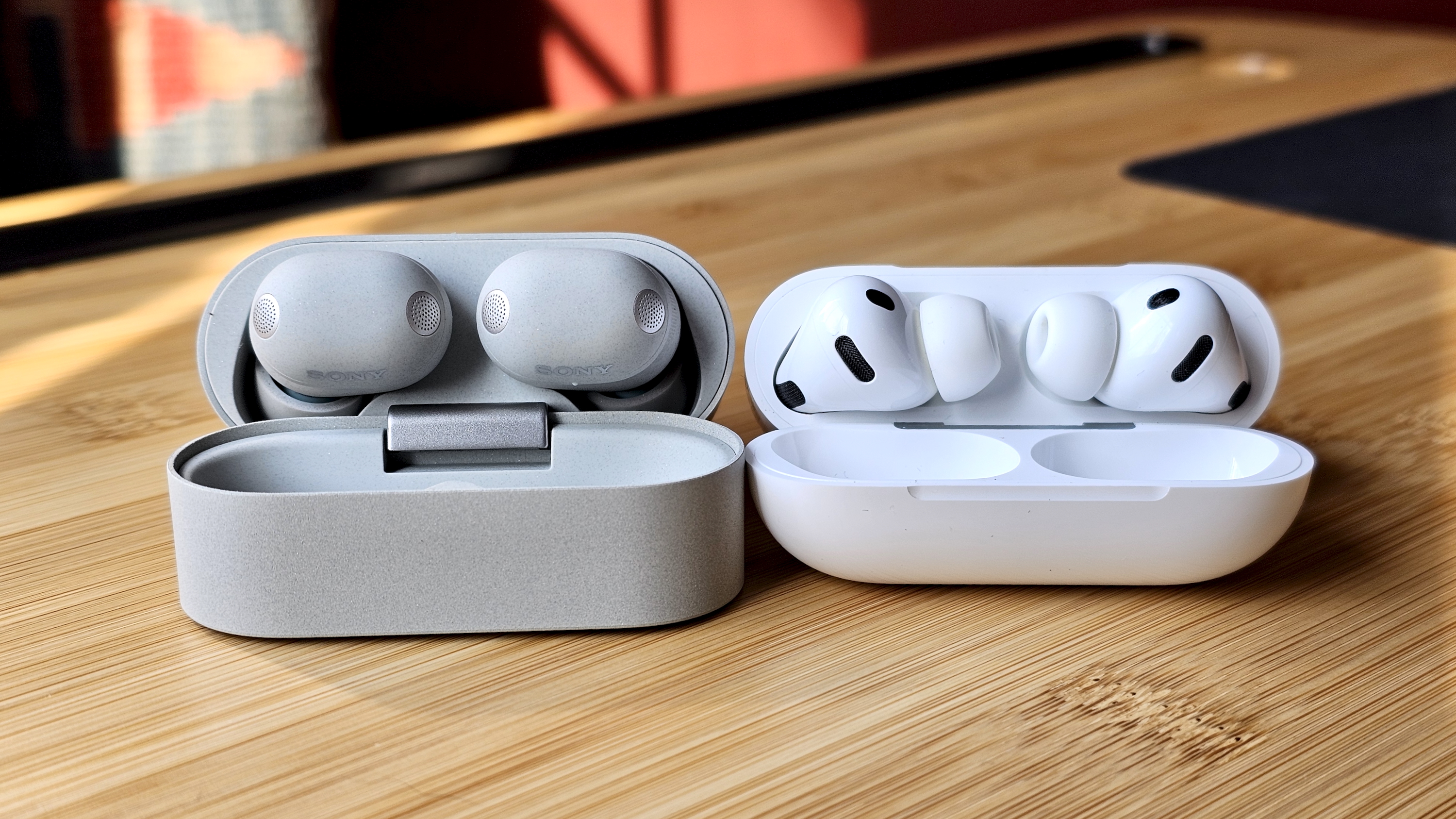 Sony WF-1000XM6 earbuds next to the Apple AirPods Pro 3, on a wooden table, in a versus-style format