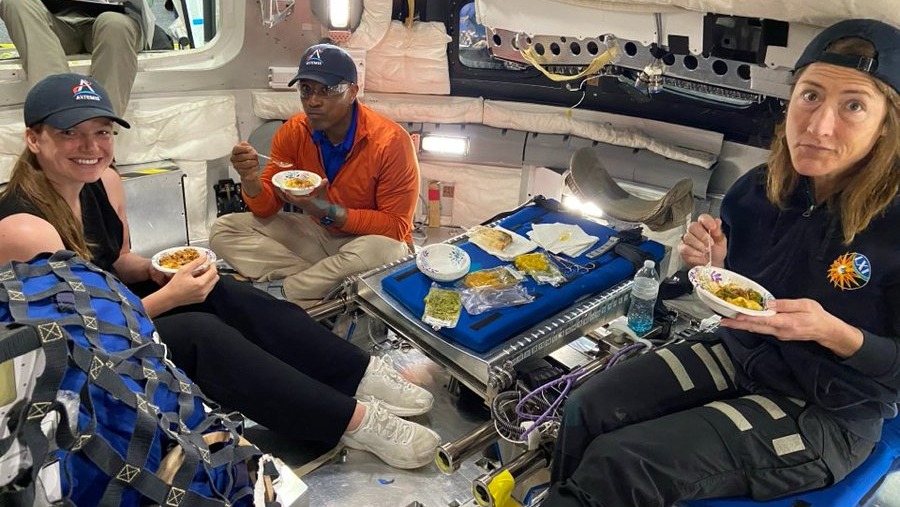 several astronauts sitting in a spacecraft with a tray of food, eating